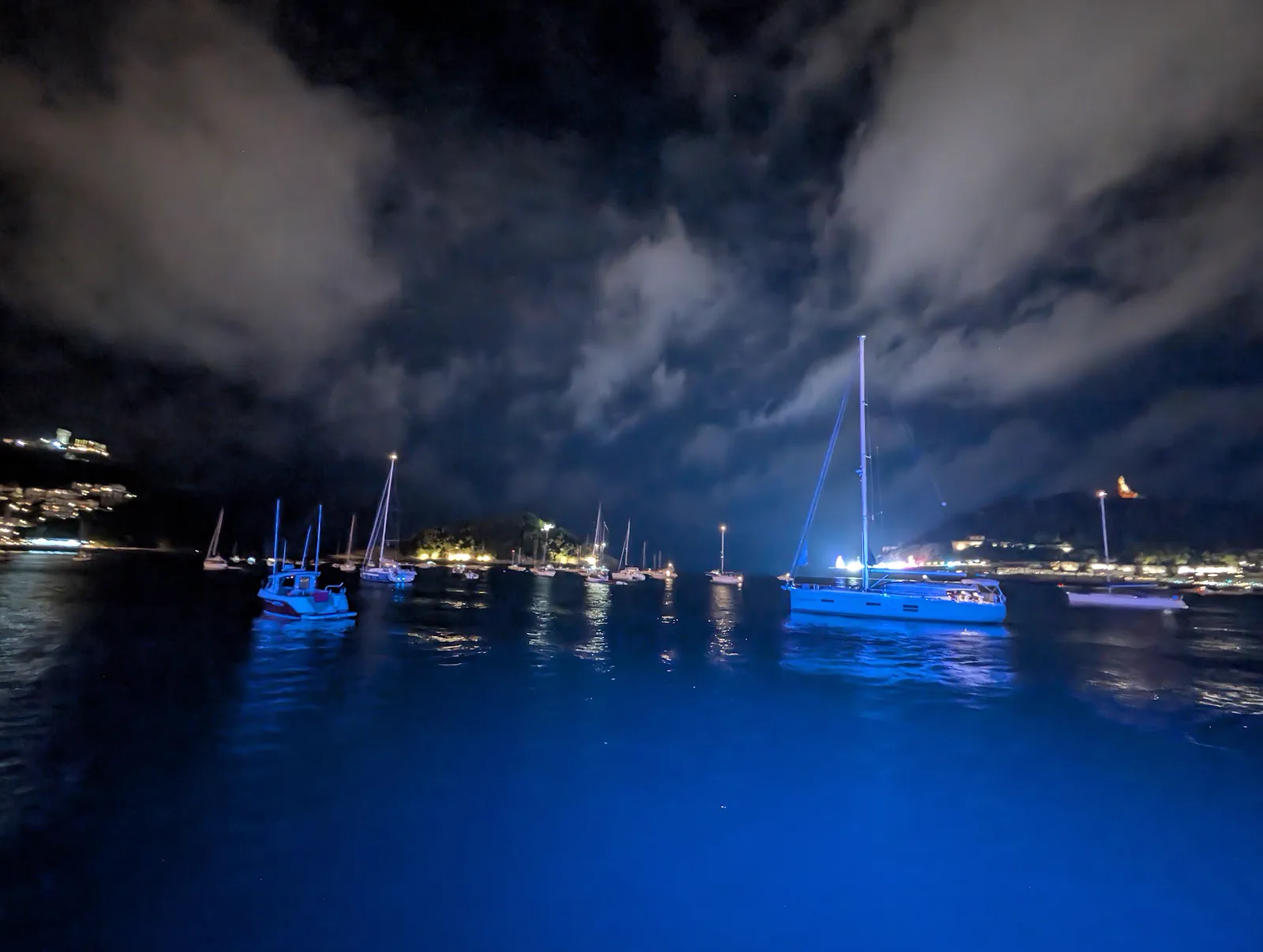 Veleros fondeados en la bahía de la Concha bajo un cielo de nubes bajas, con el agua teñida de azul por las luces del barco