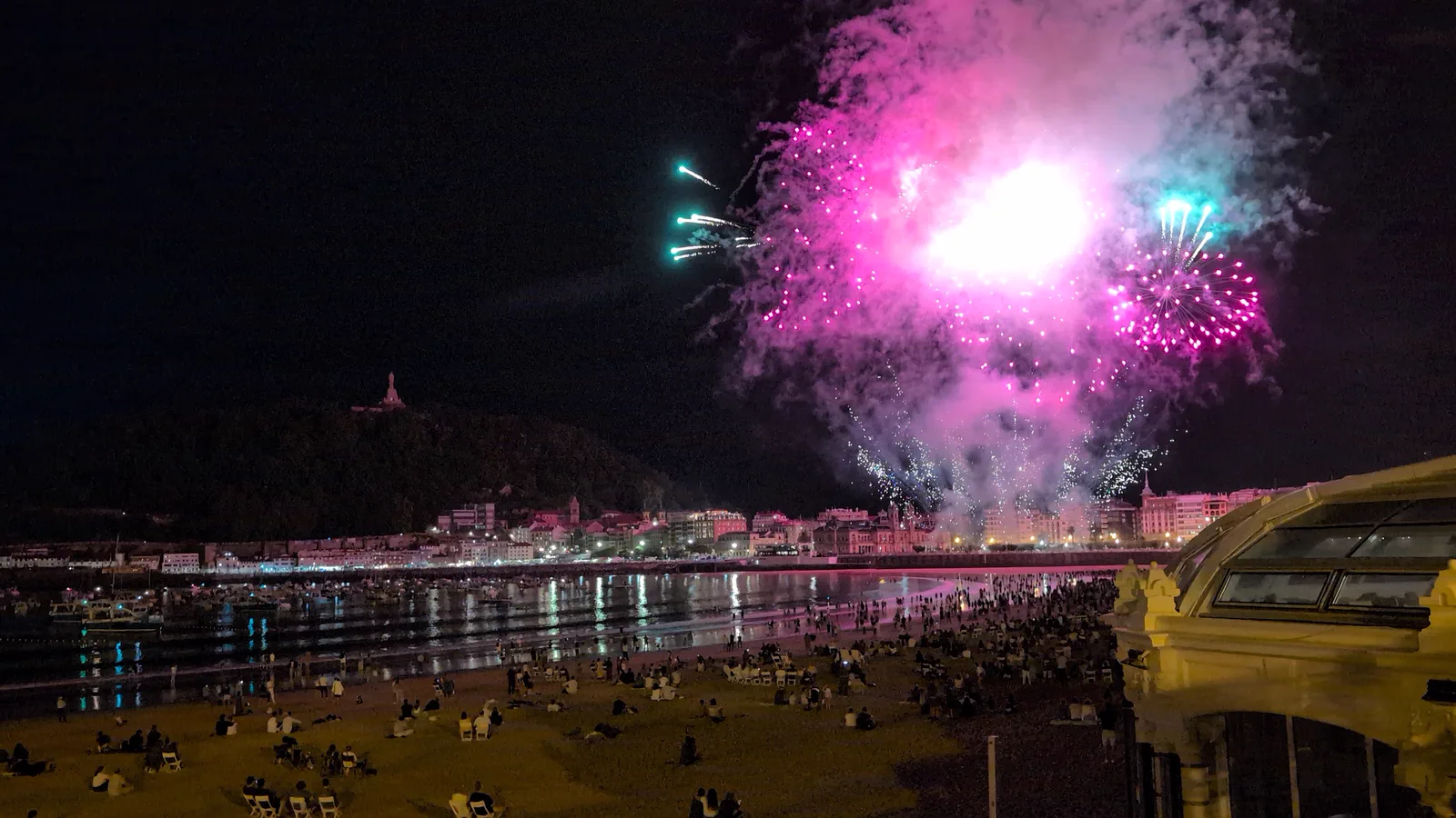 Estallido rosa y turquesa de Pirotecnia Tamarit sobre Donostia, con la playa de la Concha llena de gente en primer plano