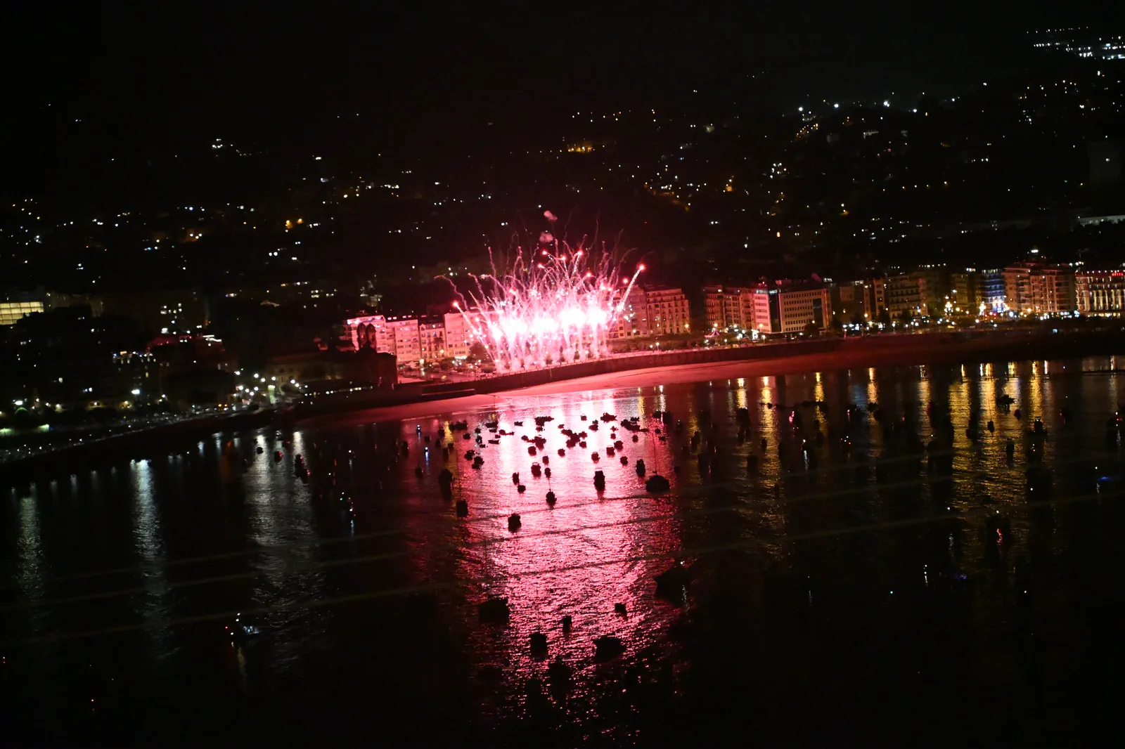 Primeros disparos rojos de Phoenix Fireworks abriéndose sobre el centro de Donostia, con el reflejo cayendo sobre el agua de la bahía