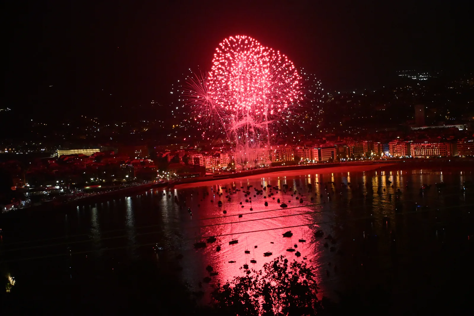 Gran palmera roja de Phoenix Fireworks sobre la bahía de la Concha vista desde el monte Igueldo, con los barcos fondeados recortados sobre el reflejo