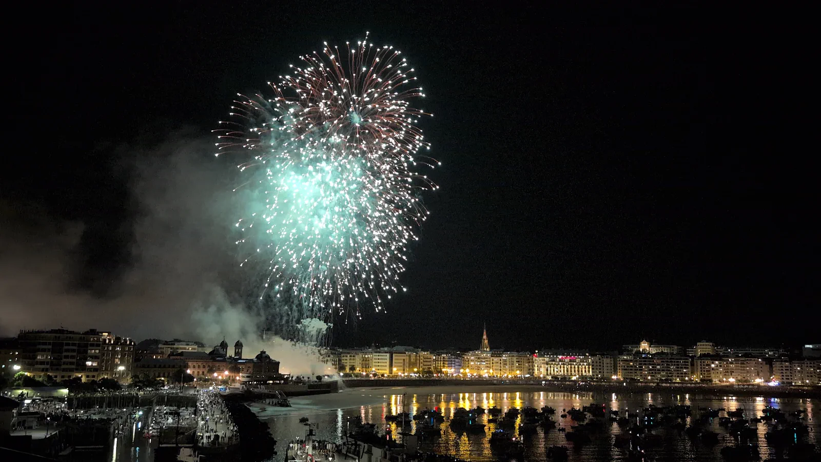 Palmera de fuegos artificiales verde y blanca abriéndose sobre la bahía de la Concha, con las luces del paseo reflejadas en el agua