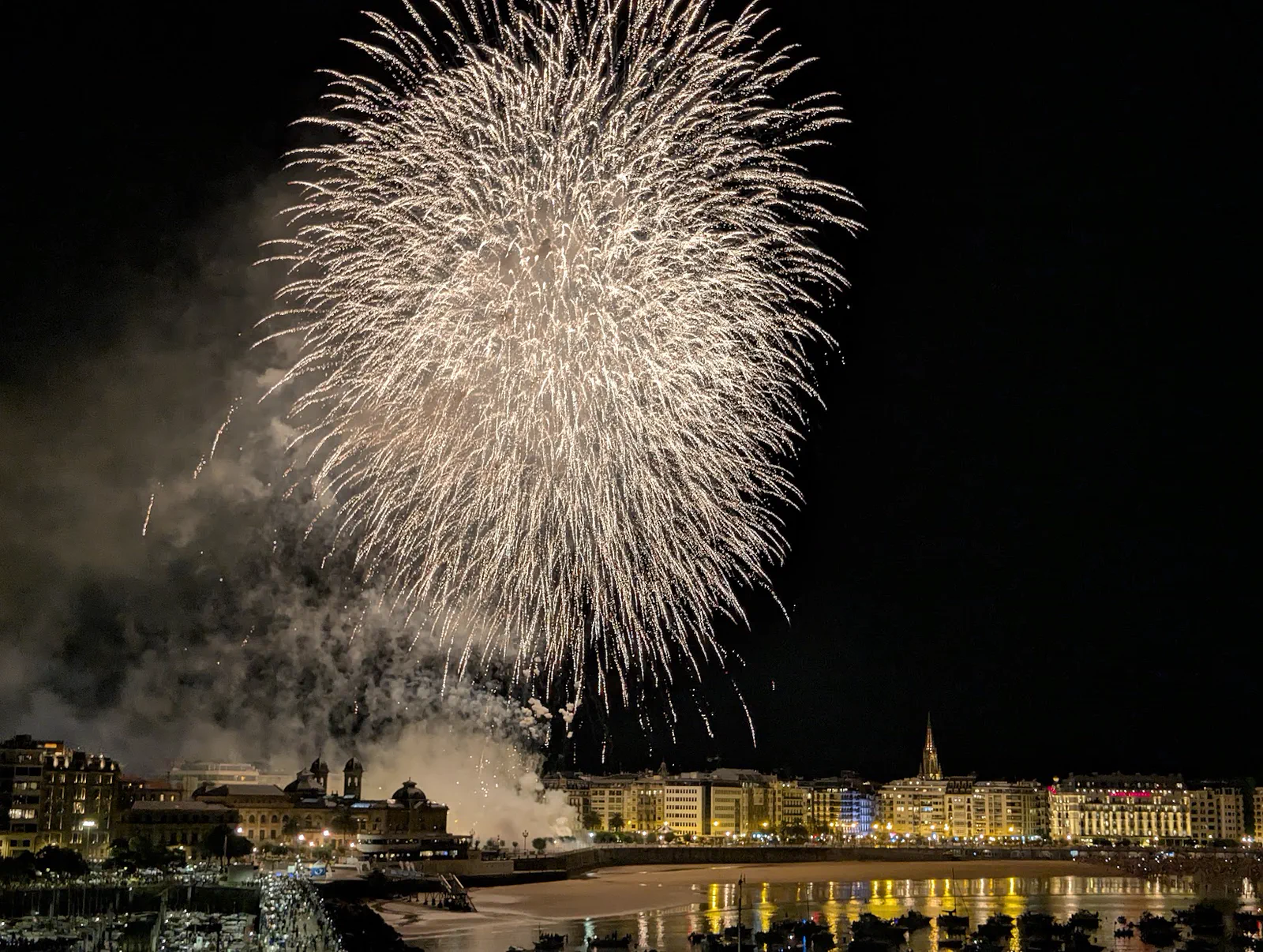 Gran esfera dorada de fuegos artificiales abriéndose sobre el centro de Donostia, con el humo acumulado a la izquierda y la playa iluminada abajo