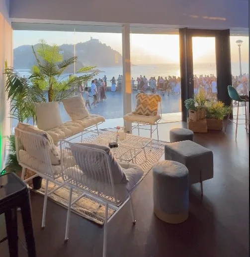 Sala Thalassa del Aquarium de San Sebastián, con sofás y ventanales abiertos sobre la playa de la Concha al atardecer
