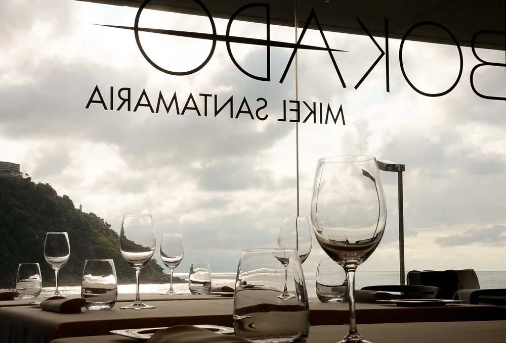 Mesa vestida junto al ventanal de Bokado Mikel Santamaría, con copas y el logo del restaurante grabado en el cristal, y la bahía de la Concha al fondo