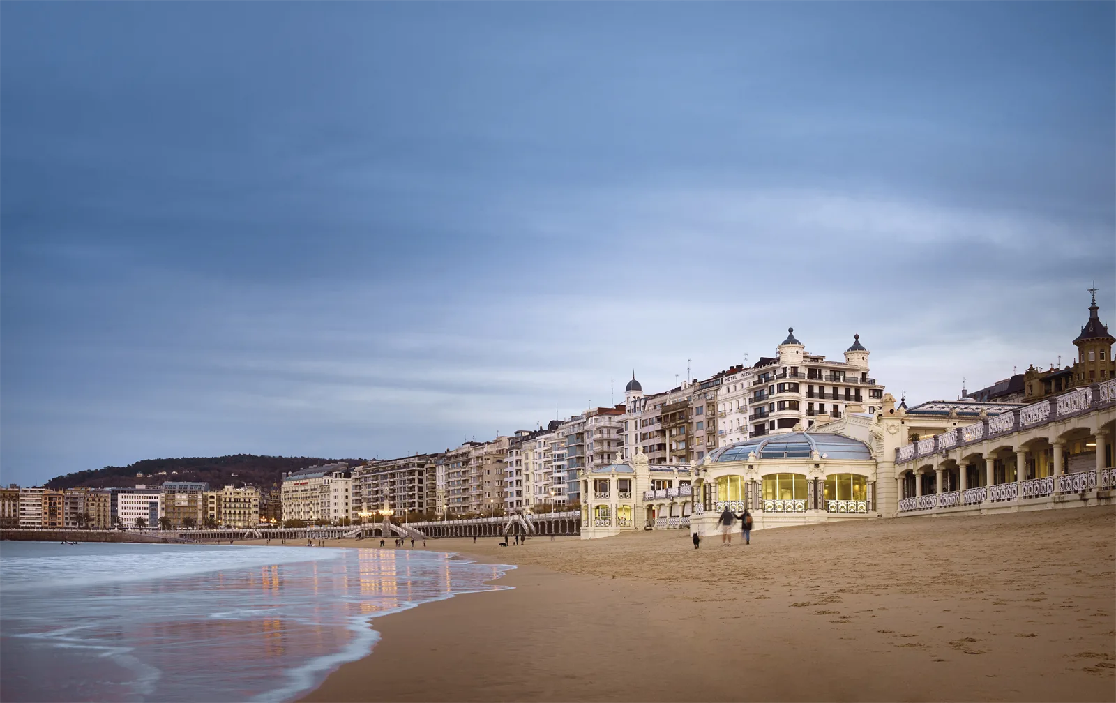 El edificio de La Perla iluminado al anochecer, sobre la arena de la playa de la Concha, con el paseo y la ciudad al fondo