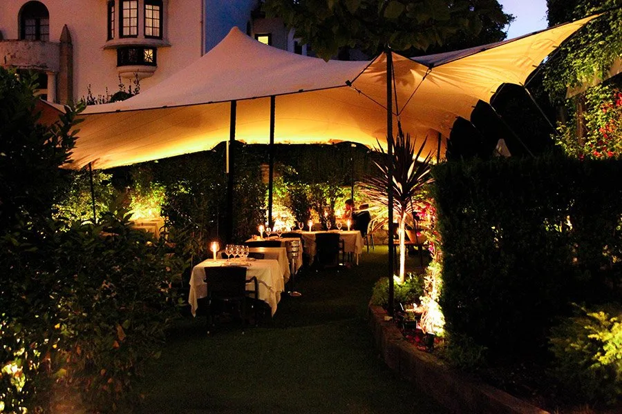 Terraza ajardinada de Saltxipi de noche, con mesas vestidas bajo una carpa iluminada junto a la villa