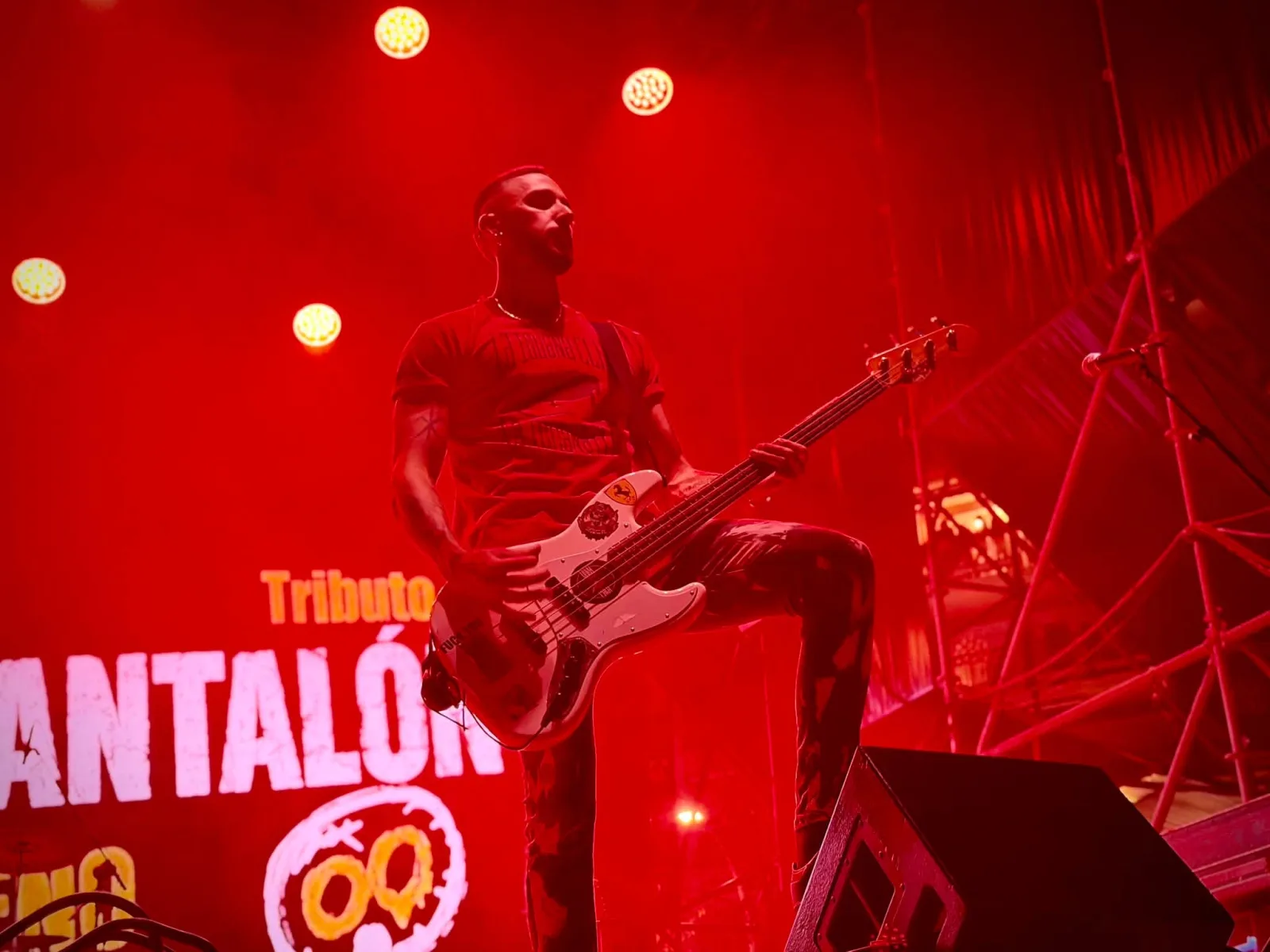 Un bajista con la pierna apoyada en un monitor toca un bajo blanco ante un photocall rojo con una calavera dibujada y las palabras «Tributo a»