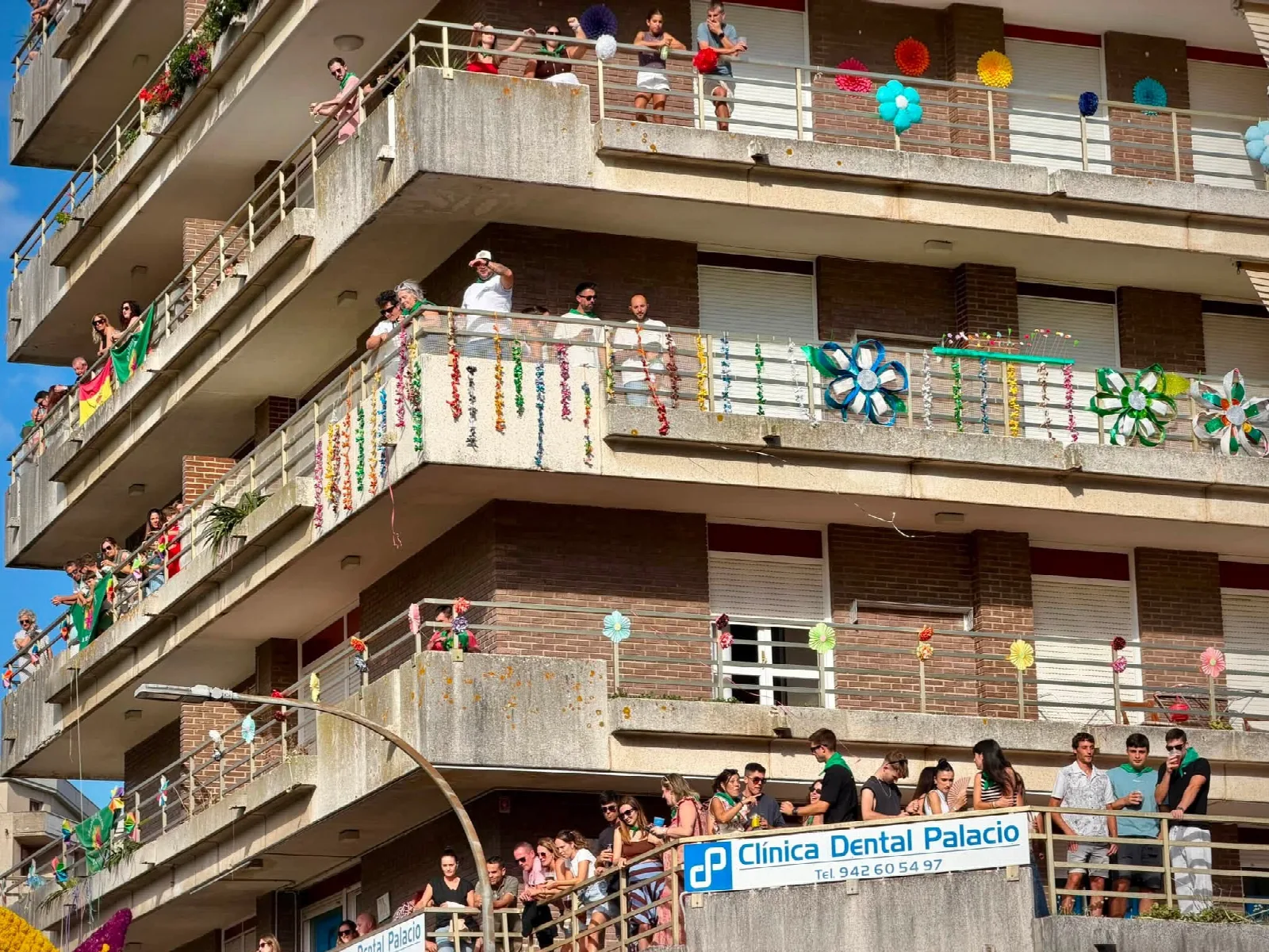 Fachada de un bloque de viviendas con varios balcones decorados con grandes flores de papel de colores y guirnaldas, y vecinos asomados mirando hacia la calle