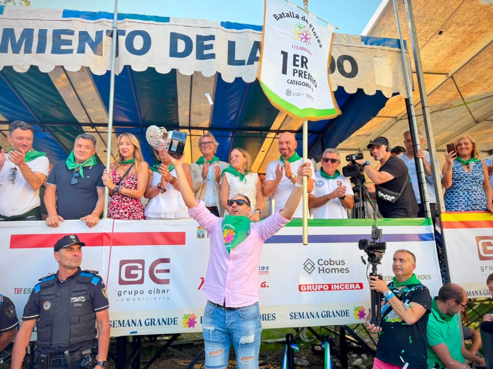 Persona con camiseta rosa y pañuelo de la Batalla de Flores levanta un trofeo y un banderín blanco con letras verdes en el que se lee «1er premio categoría A», ante la tribuna