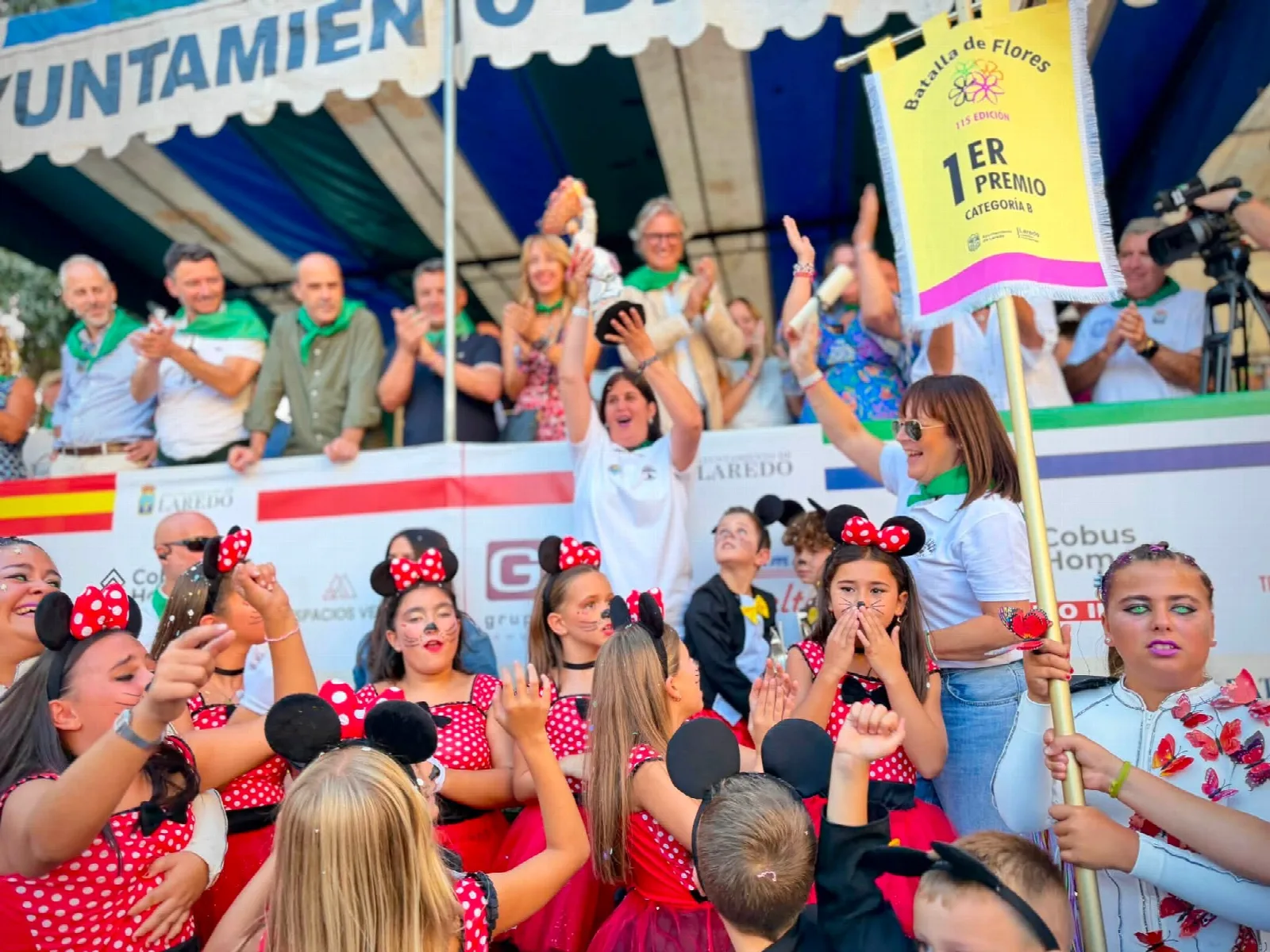 Grupo de niñas y niños disfrazados de personajes de dibujos animados celebrando bajo un banderín amarillo con la leyenda «1er premio categoría B» que sostiene una persona desde la tribuna