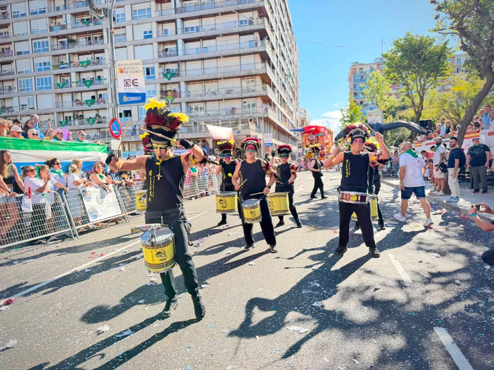 Batucada desfilando por la avenida con camisetas negras, grandes tocados de plumas rojas y amarillas y tambores amarillos, con el suelo cubierto de confeti