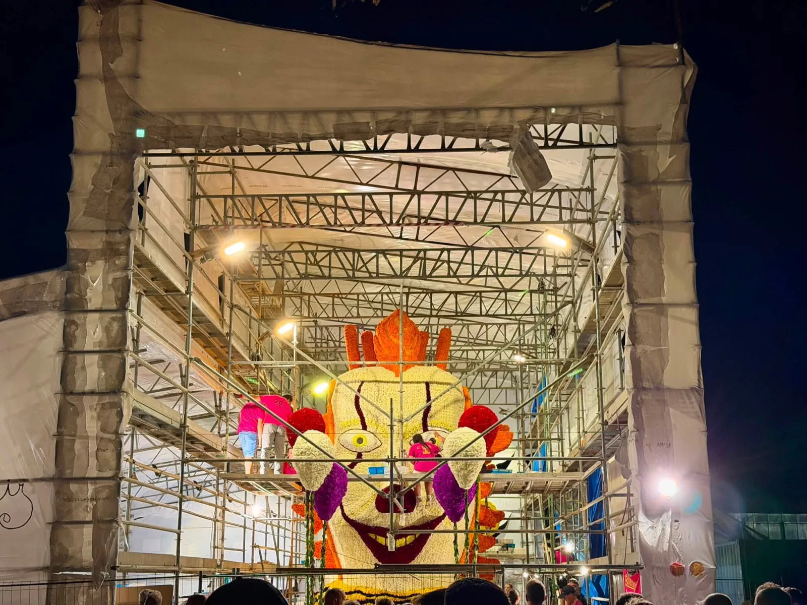 Cabeza monumental de una carroza cubierta de flor, iluminada por focos dentro de la carpa, con dos personas trabajando sobre ella