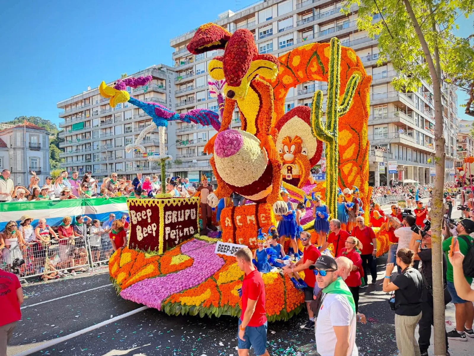 Carroza de flor con las figuras de un coyote y un correcaminos de dibujos animados, con la placa del título en la parte inferior y público tras las vallas