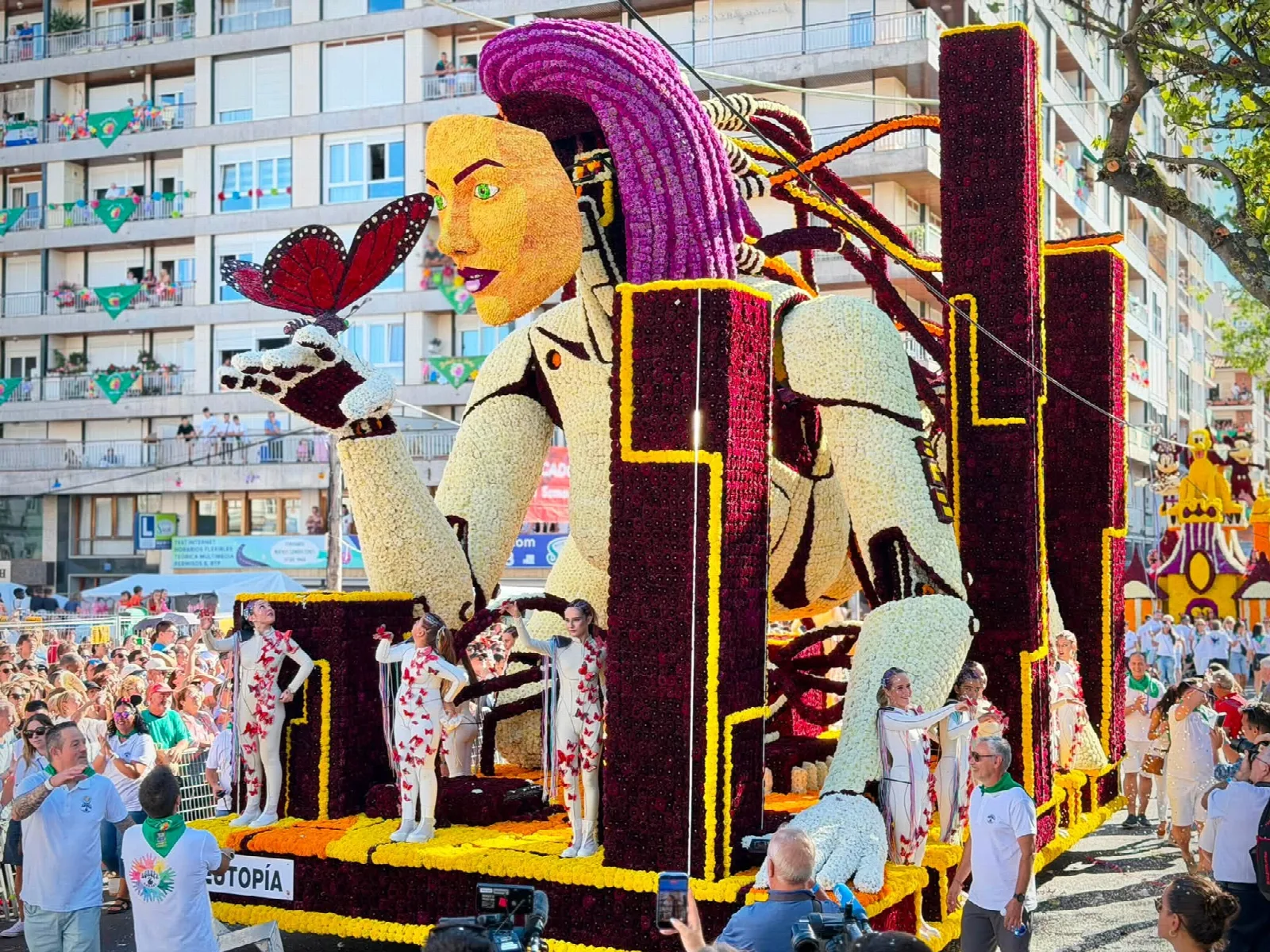 Carroza de flor con una figura femenina monumental de bata blanca y pelo morado que sostiene una gran mariposa, con la placa del título en la parte inferior y público al fondo