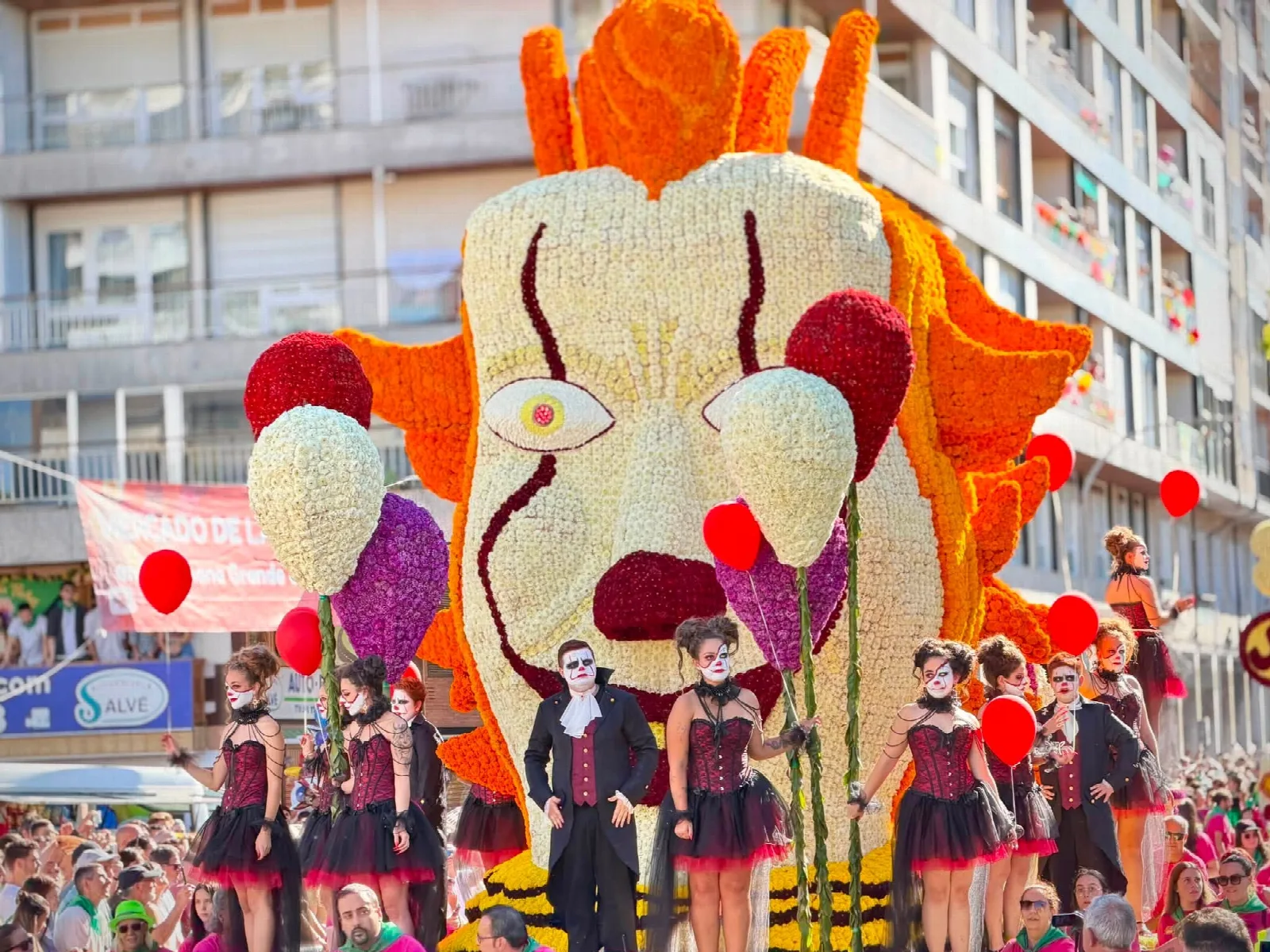 Carroza de flor con un payaso monumental de gesto inquietante y varios globos rojos de gran tamaño, avanzando entre el público