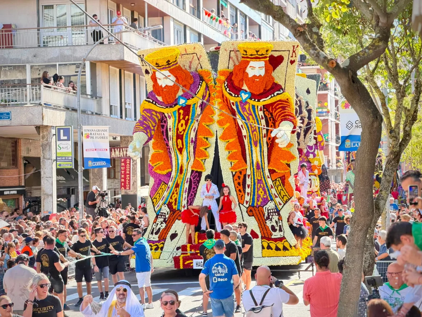 Carroza monumental de flor natural con dos reyes de la baraja de gran tamaño avanzando por una avenida flanqueada por miles de personas, con la placa del título en la parte inferior