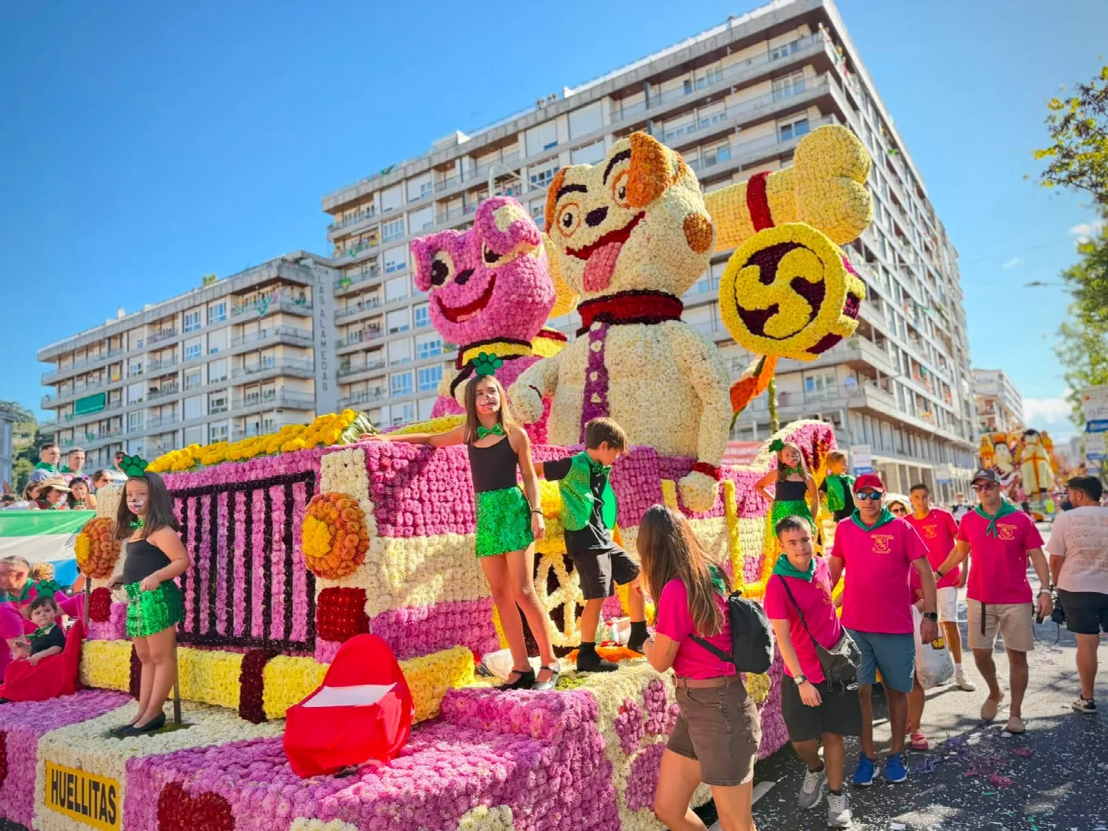 Carroza de flor rosa y amarilla con dos figuras de perro y un hueso de gran tamaño, con niños disfrazados sobre la plataforma y la placa del título delante