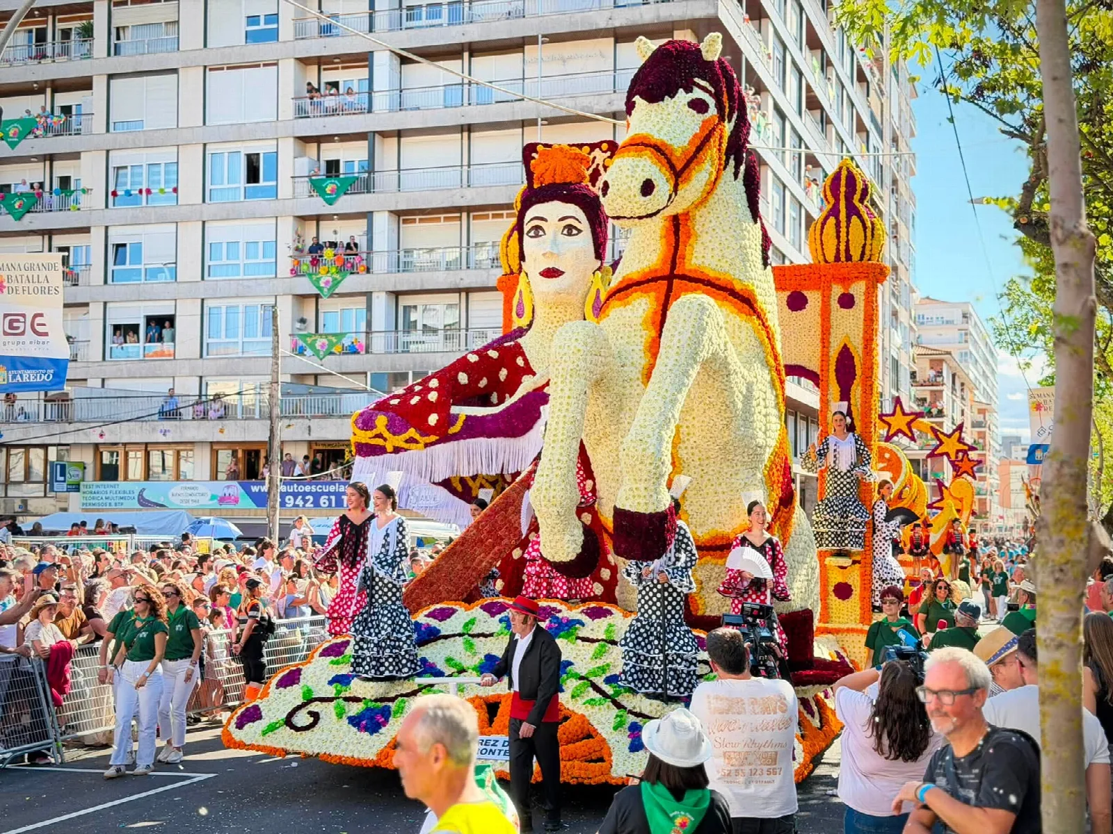 Carroza de flor con un caballo andaluz de gran tamaño y motivos de temática jerezana, con personas vestidas de flamenca sobre la plataforma y la placa del título delante