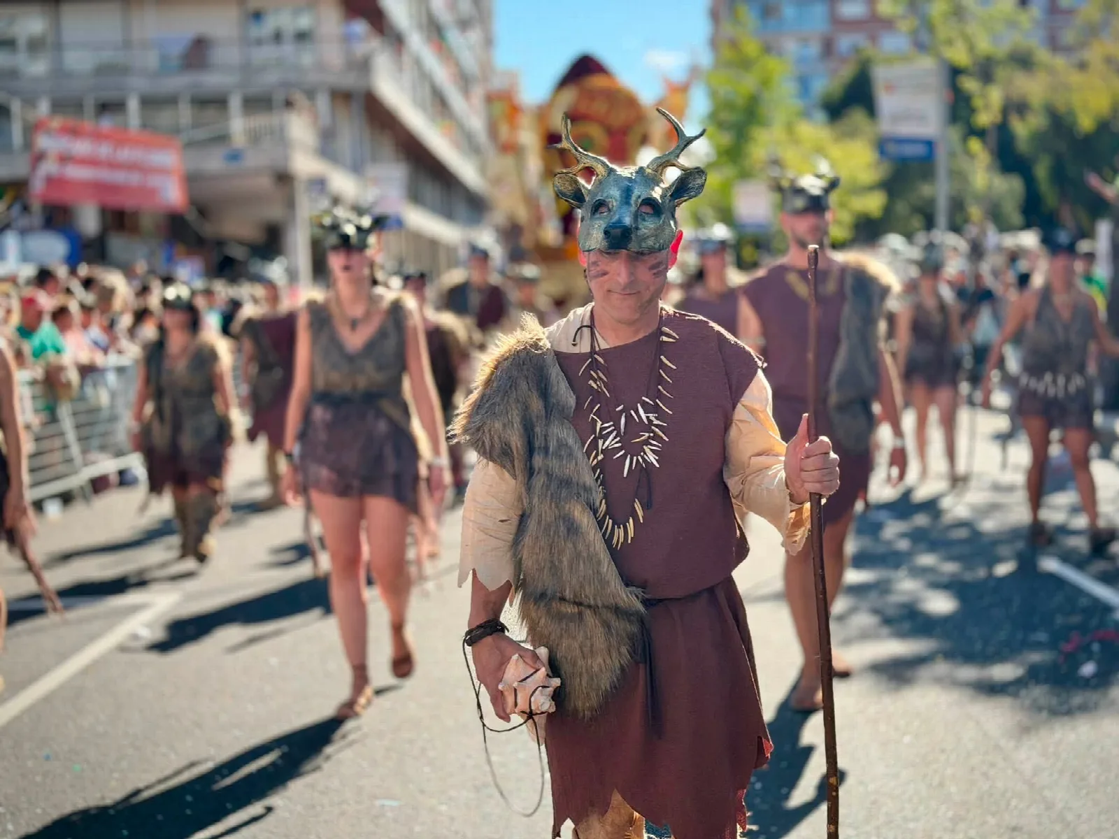 Grupo de personas desfilando a pie con vestuario de pieles, tocados de asta y máscaras de cráneo de ciervo, entre el público que las mira desde las vallas