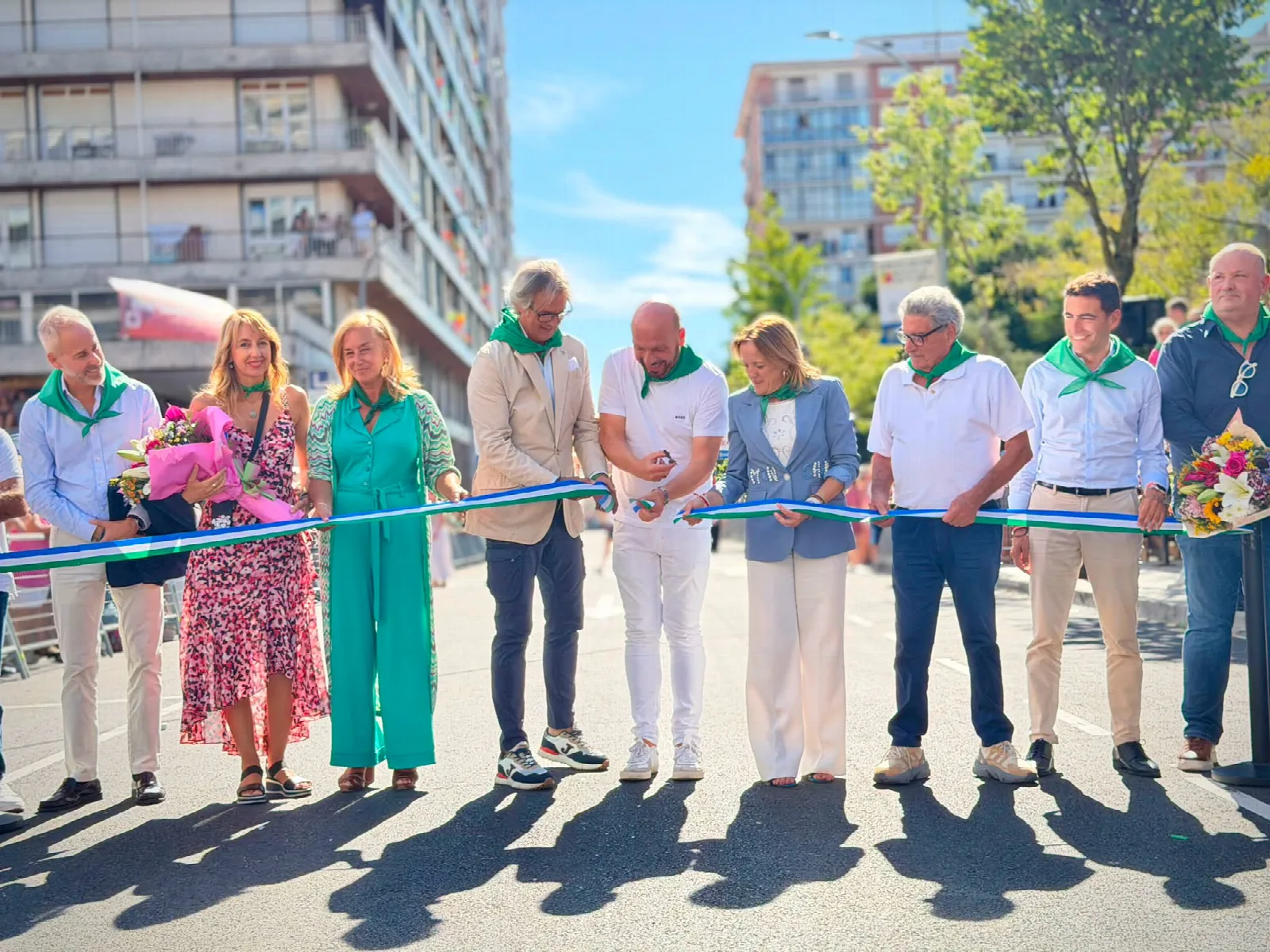 Una decena de personas con pañuelo verde de fiestas sostienen una cinta verde, blanca y azul que cruza la calzada mientras dos de ellas la cortan, con bloques de viviendas engalanados al fondo