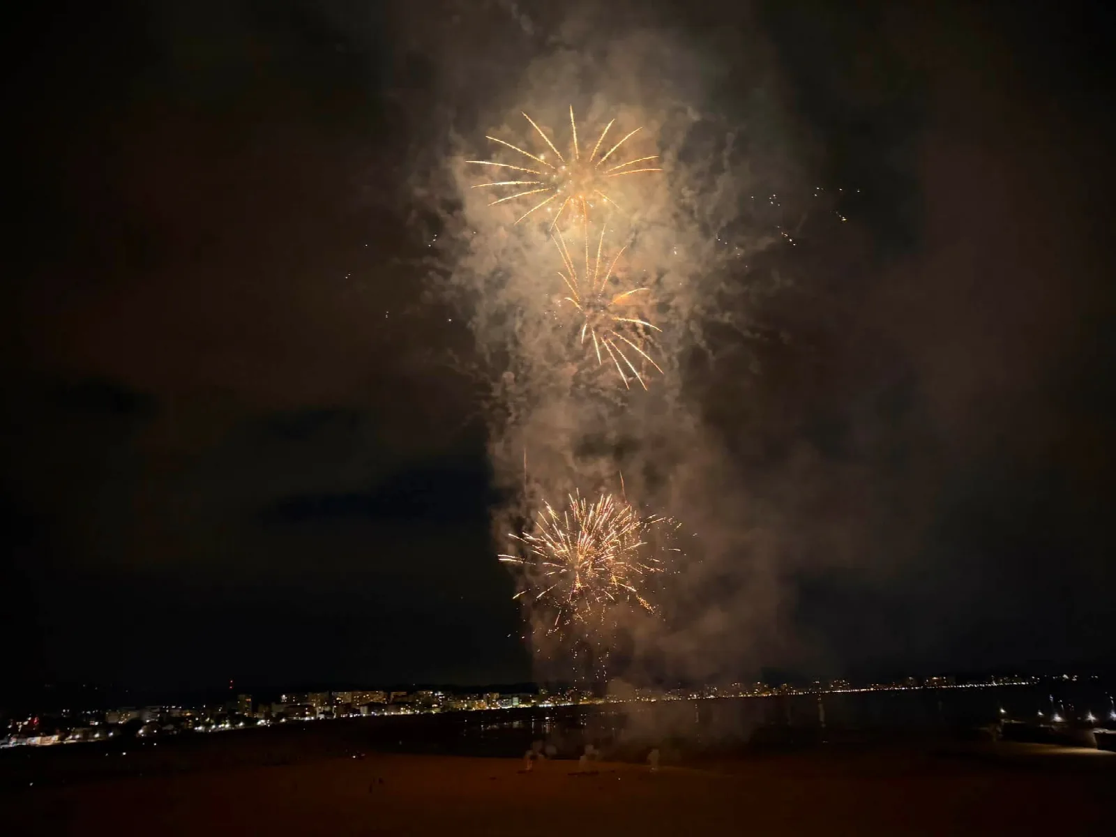 Tres explosiones de fuegos artificiales doradas sobre la playa a oscuras, con la línea de luces del pueblo y la ría al fondo