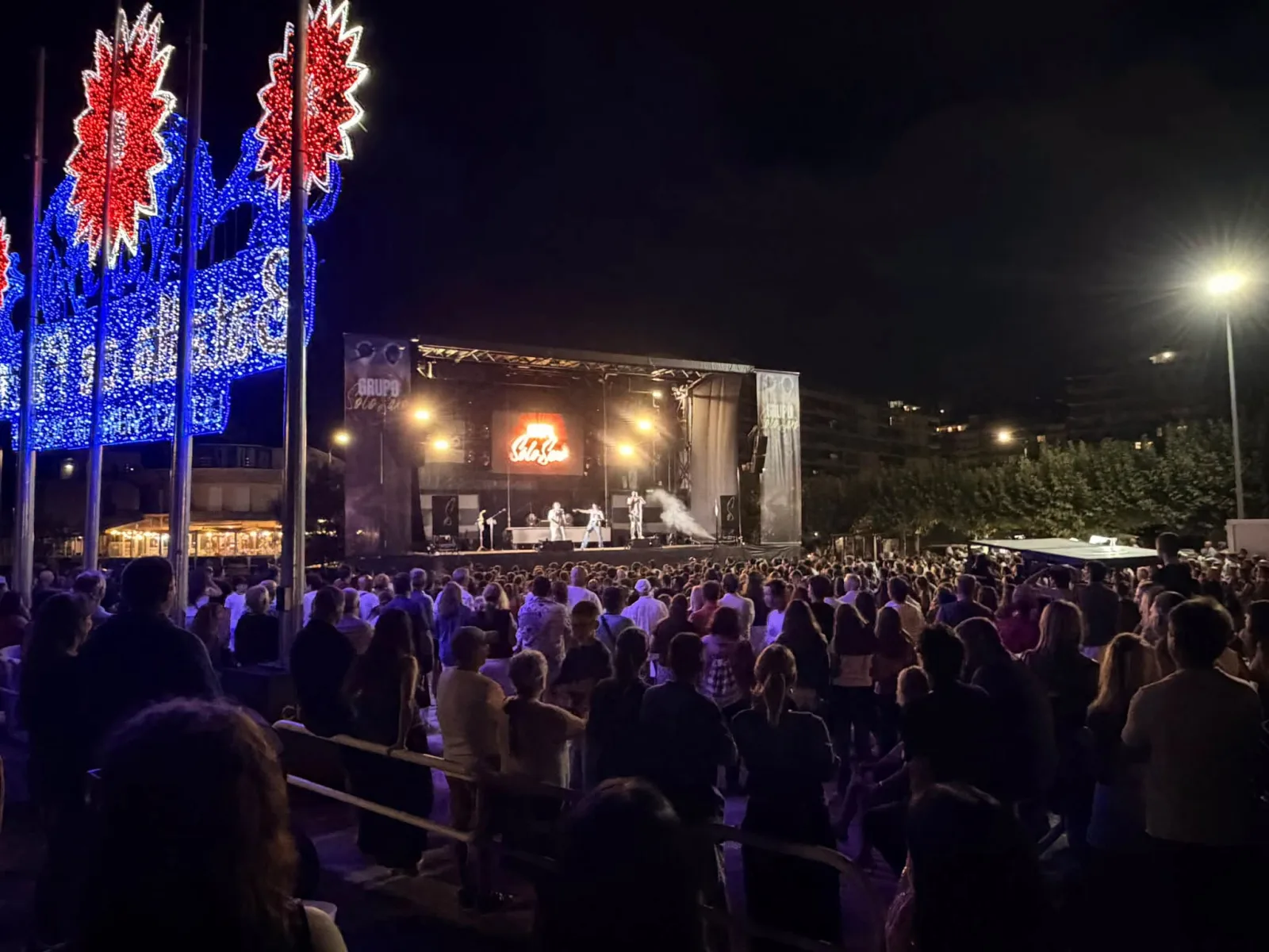 Escenario con el rótulo del Grupo Solo Saxo y varios músicos actuando, visto desde el fondo de una explanada llena de gente de pie, con los arcos de luces de fiestas en rojo y azul a la izquierda