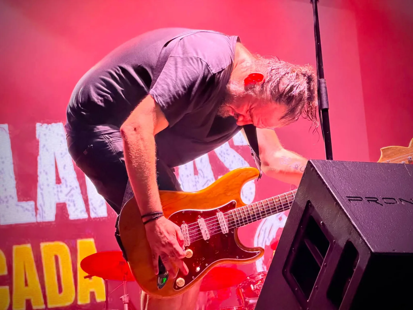 Un guitarrista se dobla sobre una guitarra naranja para manipular los mandos, con el pelo empapado y un photocall rojo detrás