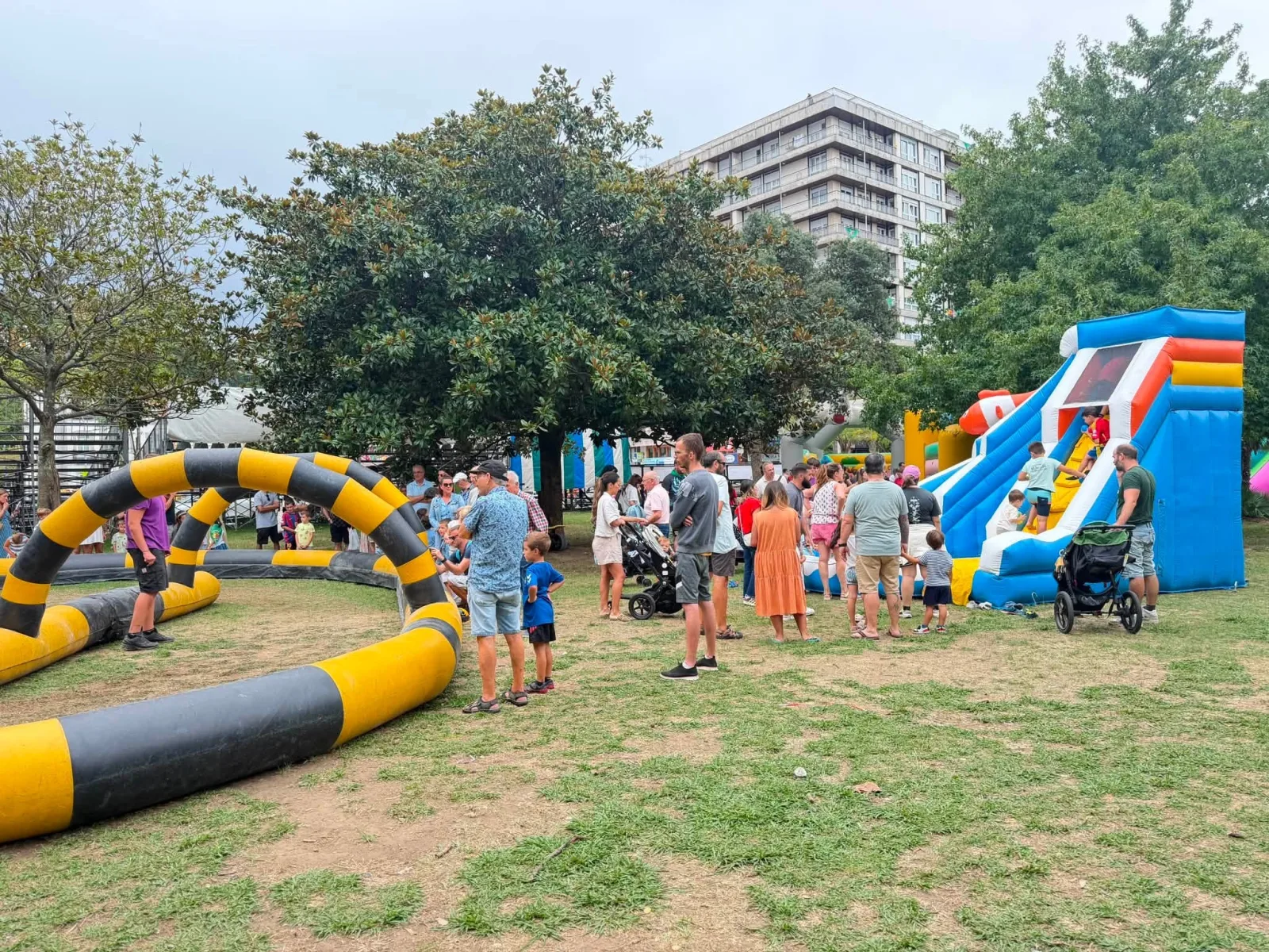Zona infantil al aire libre con un circuito hinchable de obstáculos amarillo y negro en primer término y un tobogán hinchable azul y rojo a la derecha, rodeados de familias con niños y carritos de bebé bajo los árboles de la Alameda