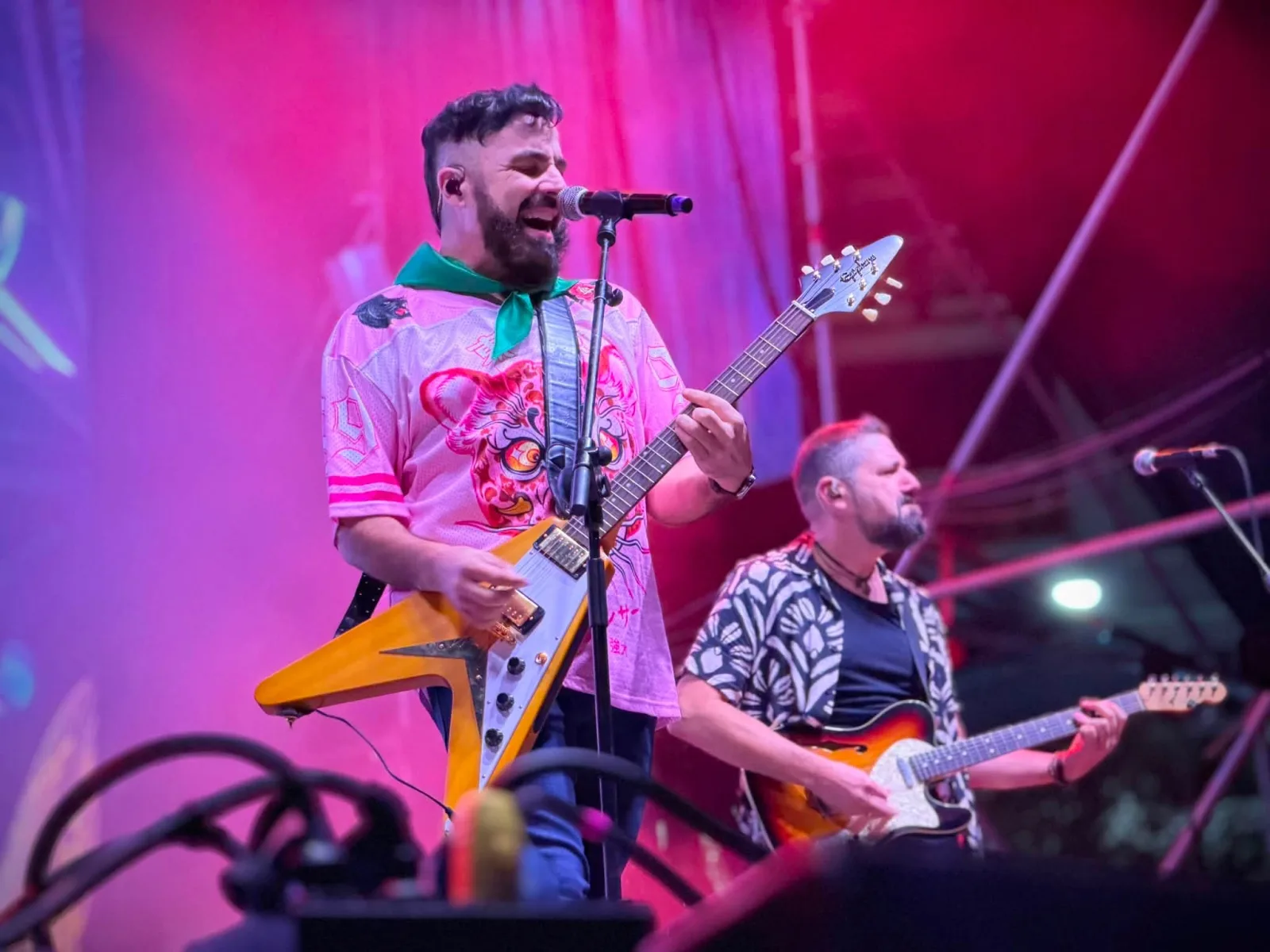 Huecco cantando ante el micrófono con una guitarra eléctrica de cuerpo triangular, camiseta rosa con un tigre estampado y pañuelo verde al cuello, con un segundo guitarrista a su derecha y el escenario iluminado en rosa y rojo