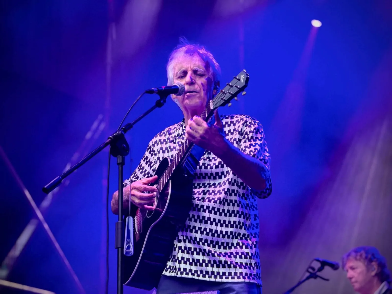 Primer plano de Kiko Veneno cantando ante el micrófono con los ojos cerrados y una guitarra acústica, con camisa de estampado geométrico en blanco y negro y fondo de luces azules