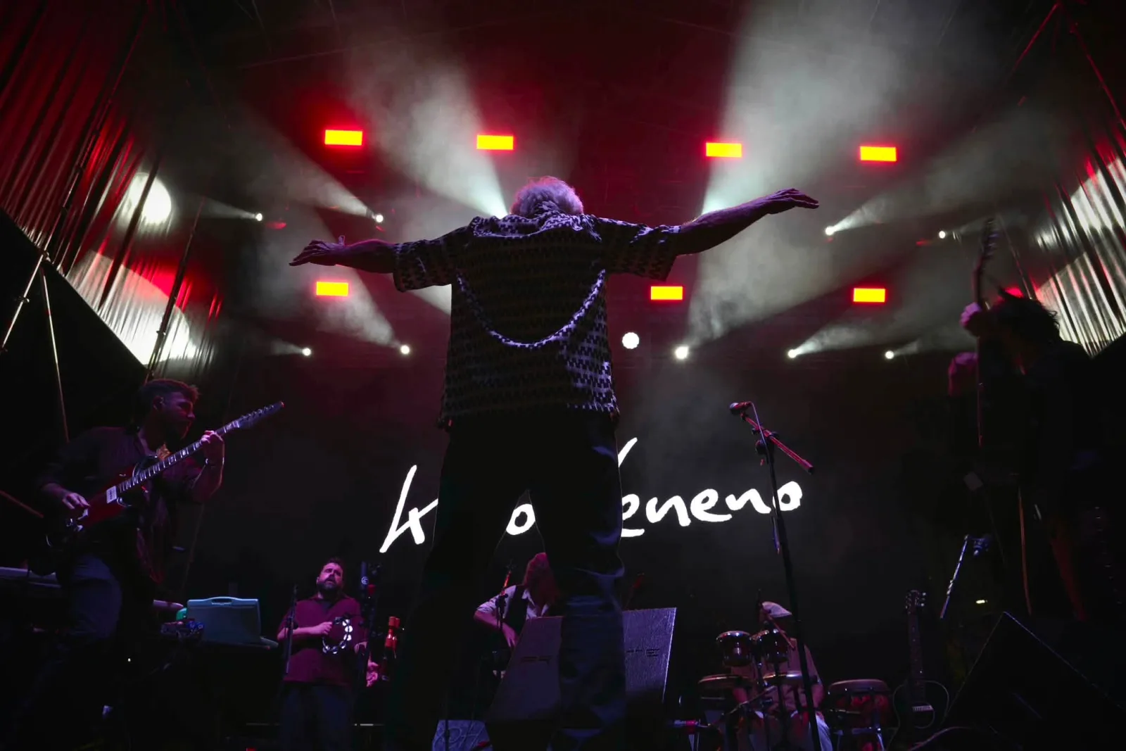 Kiko Veneno de espaldas al fotógrafo, con los brazos completamente abiertos, ante un escenario lleno de humo iluminado en rojo y su nombre escrito a mano proyectado en la pantalla del fondo