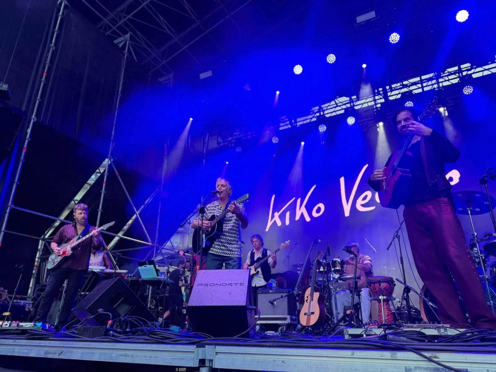 Kiko Veneno cantando ante un micrófono con una guitarra pequeña en el centro de un escenario iluminado en azul, acompañado por un guitarrista eléctrico, un bajista, un percusionista y un guitarrista con guitarra española