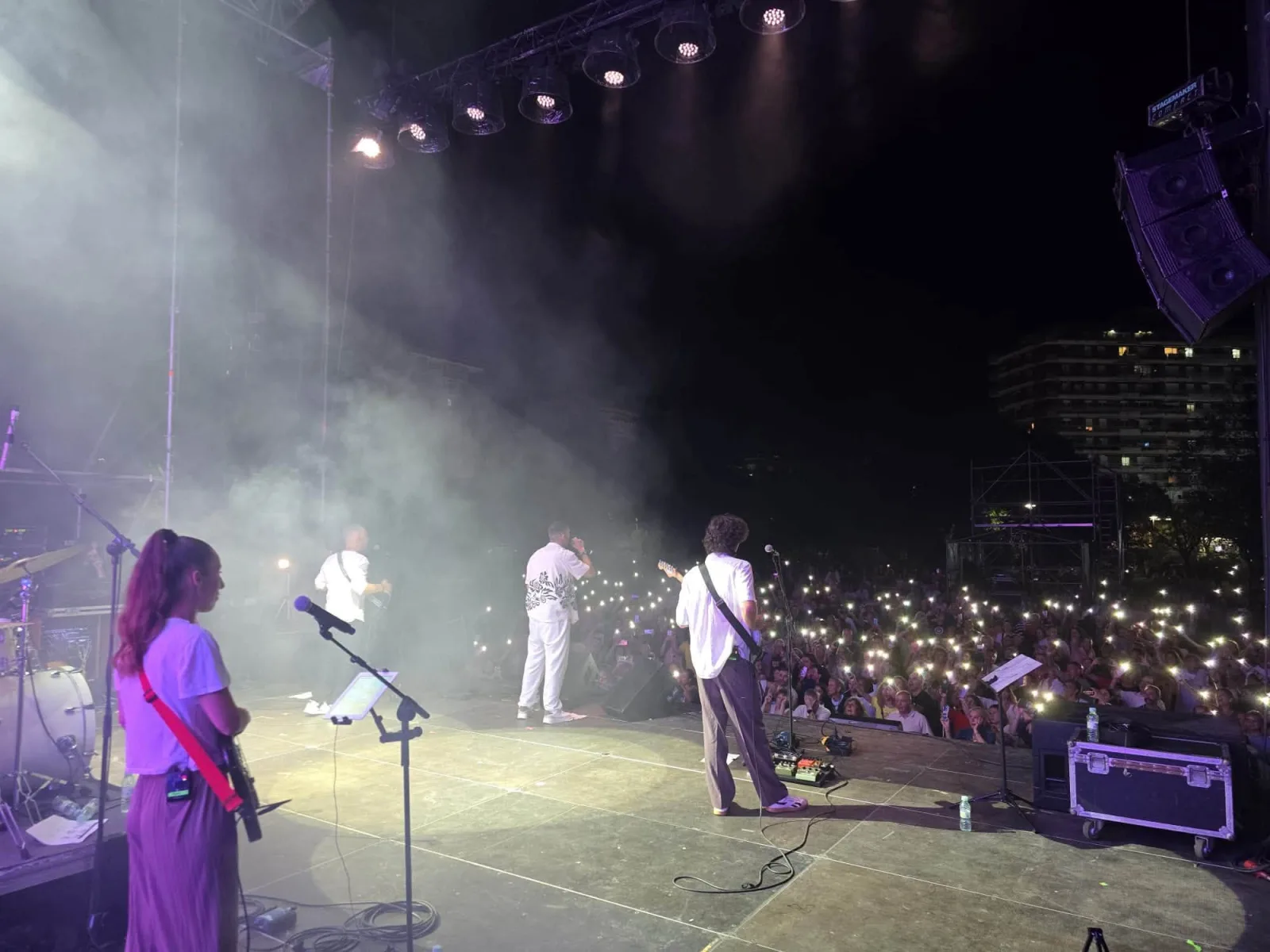 Escena nocturna tomada desde un lateral del escenario, entre el humo de los focos: el cantante de blanco y dos músicos de espaldas y, frente a ellos, cientos de linternas de móvil encendidas entre el público