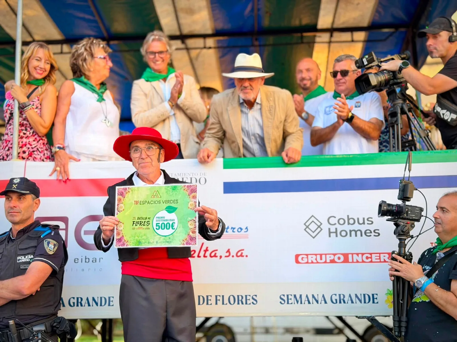 Persona con sombrero rojo sostiene ante la tribuna un diploma con fondo de flores en el que se lee «TBF Espacios Verdes — 115 edición Batalla de Flores — Premio Sostenibilidad 500 €»