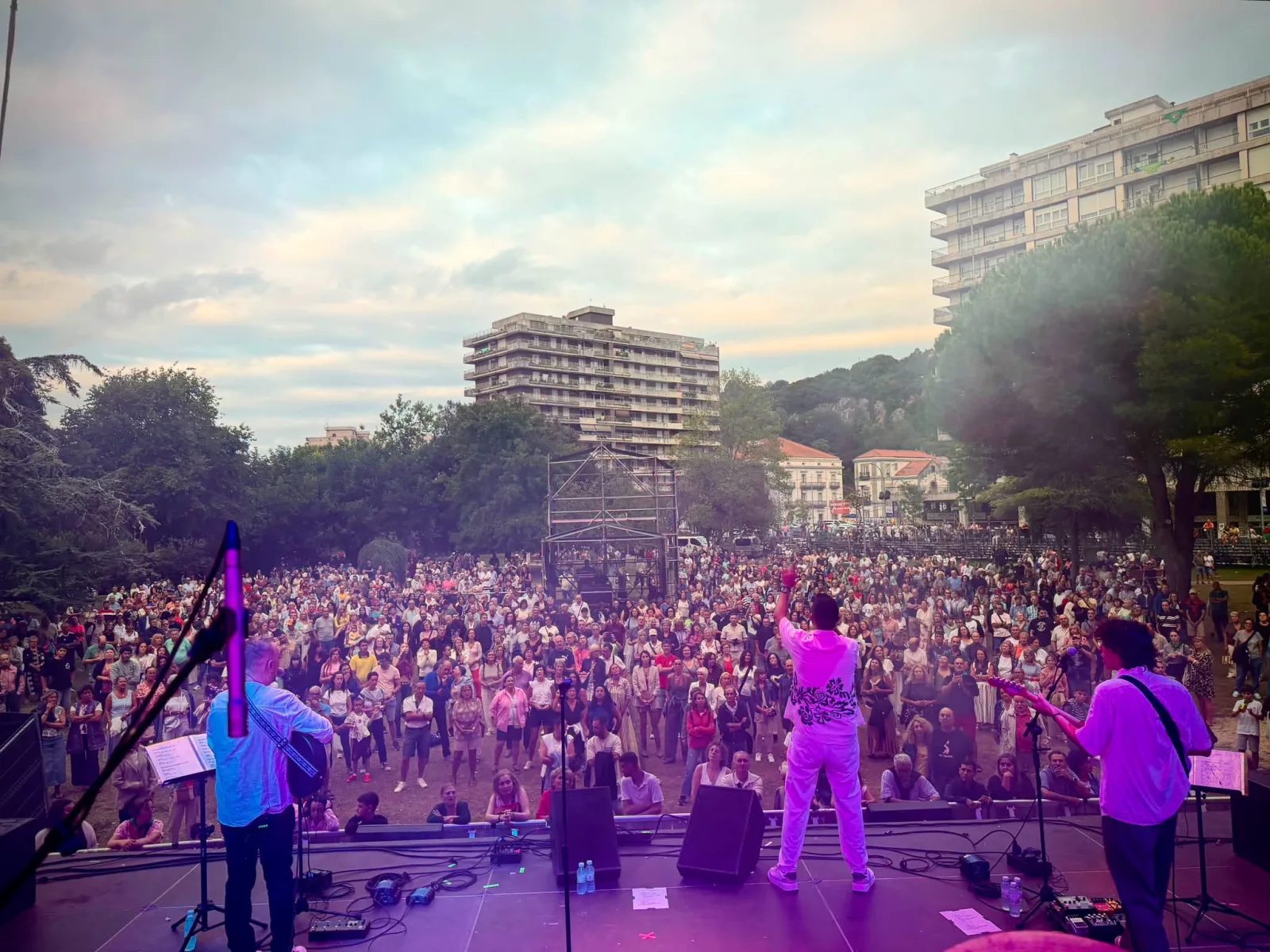Fotografía tomada desde el escenario, de espaldas a los músicos: el cantante vestido de blanco con el brazo en alto en el centro, dos guitarristas a los lados bajo luz violeta y, delante, la explanada de la Alameda Miramar llena de gente bajo un cielo de atardecer