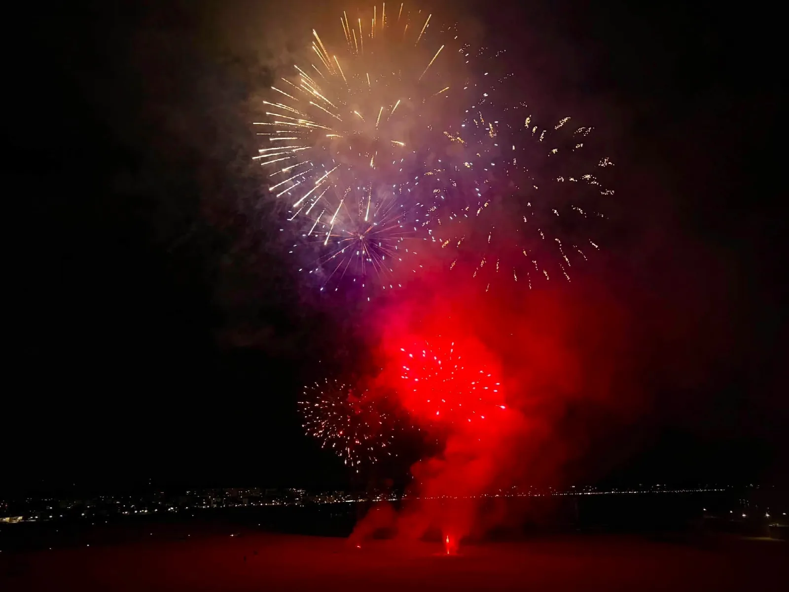 Explosiones doradas y violetas en la parte alta del cielo y una gran mancha roja de fuegos más abajo, sobre la playa y la línea de luces de la costa