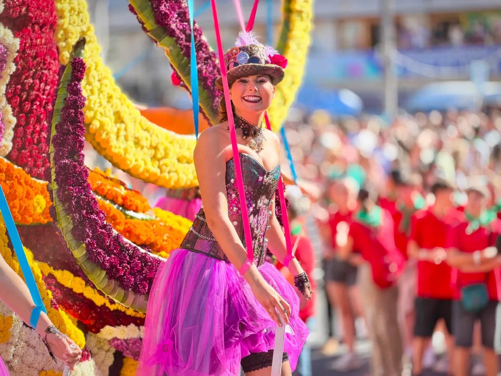 Mujer con corsé, falda de tul magenta y chistera adornada con engranajes y plumas, sujeta a unas cintas de colores junto a un lateral de carroza cubierto de flor