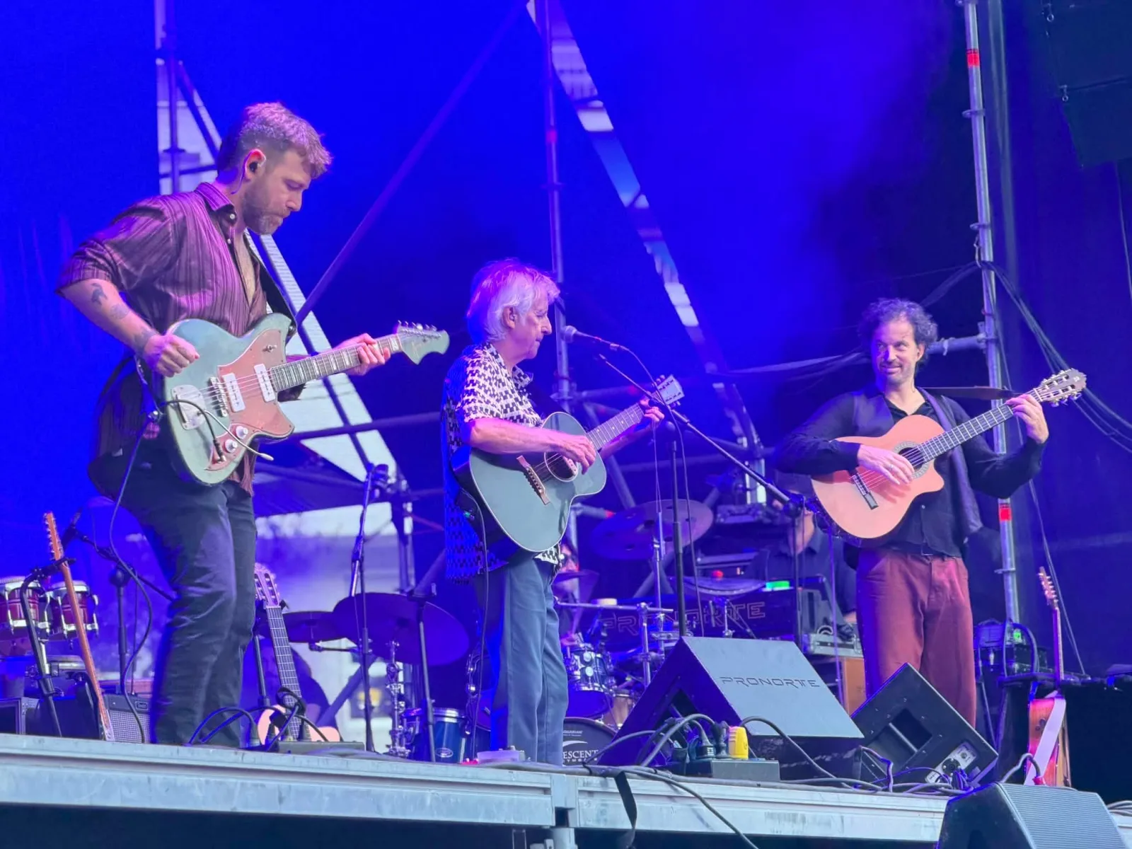 Tres músicos tocando en fila en un escenario iluminado en azul: un guitarrista eléctrico inclinado hacia delante, Kiko Veneno con guitarra acústica ante el micrófono y un tercer músico con guitarra española