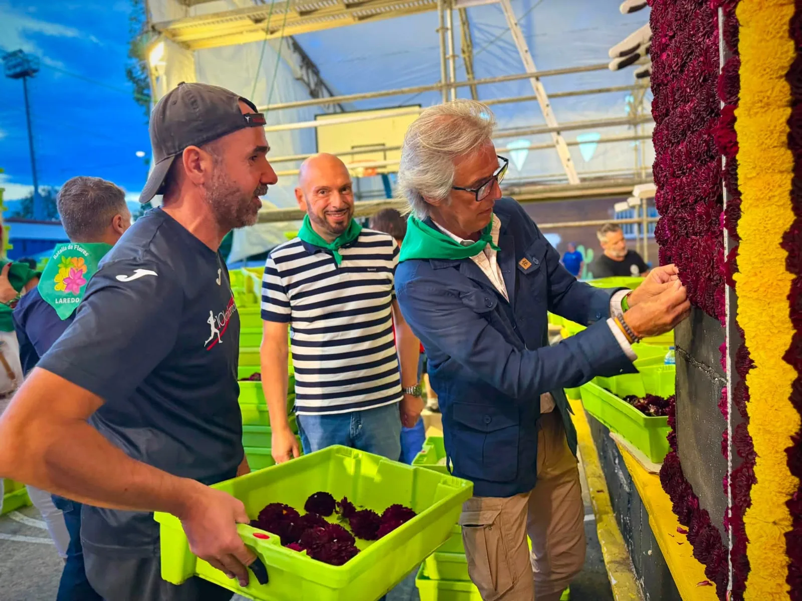 Persona con pañuelo verde de las fiestas colocando una flor granate en el lateral de una carroza mientras un carrocista sostiene la caja de flor